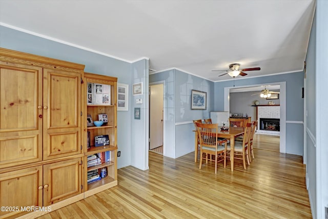 dining space featuring light wood-style flooring, a brick fireplace, crown molding, and a ceiling fan