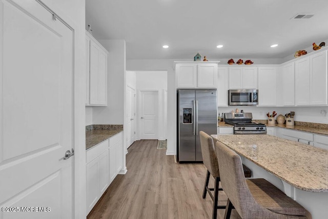 kitchen featuring light stone countertops, stainless steel appliances, white cabinetry, light wood-type flooring, and a kitchen bar