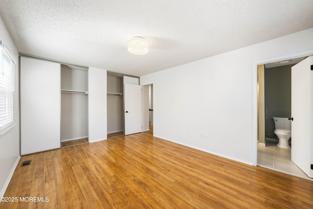 unfurnished bedroom featuring visible vents, two closets, baseboards, wood finished floors, and ensuite bath