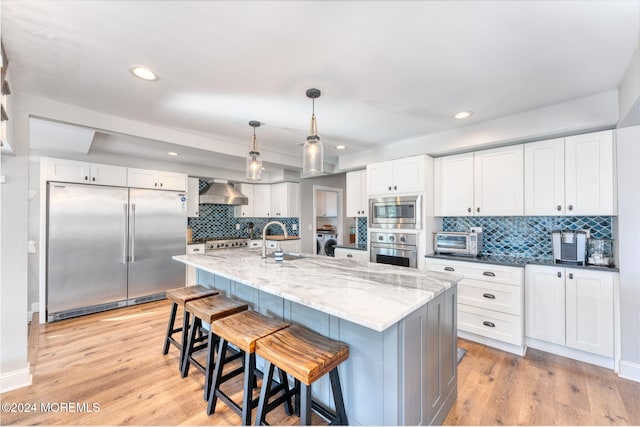 kitchen with wall chimney range hood, built in appliances, light wood-style floors, white cabinets, and a sink