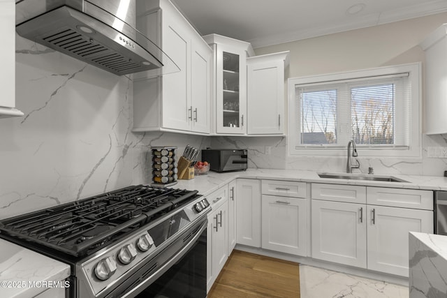 kitchen with stainless steel gas stove, a sink, wall chimney exhaust hood, white cabinets, and crown molding