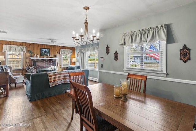 dining space featuring hardwood / wood-style floors, wood walls, a brick fireplace, ceiling fan with notable chandelier, and baseboard heating