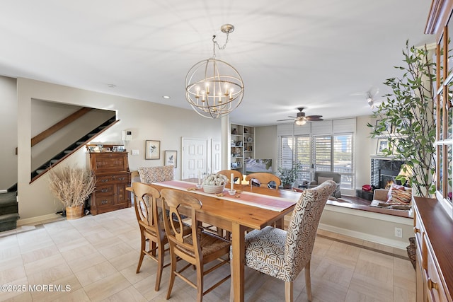 dining room featuring a glass covered fireplace, stairway, ceiling fan with notable chandelier, and baseboards