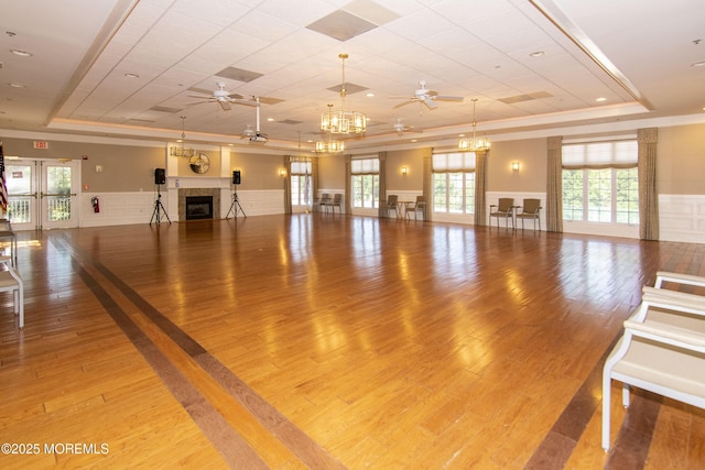 workout room featuring a raised ceiling, a fireplace, wainscoting, and ceiling fan with notable chandelier