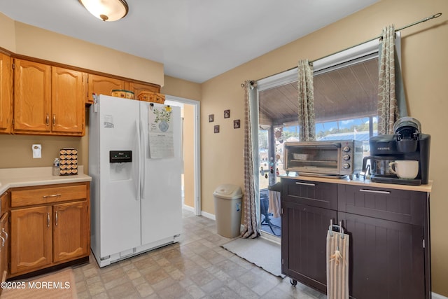 kitchen featuring brown cabinetry, white refrigerator with ice dispenser, light countertops, and baseboards