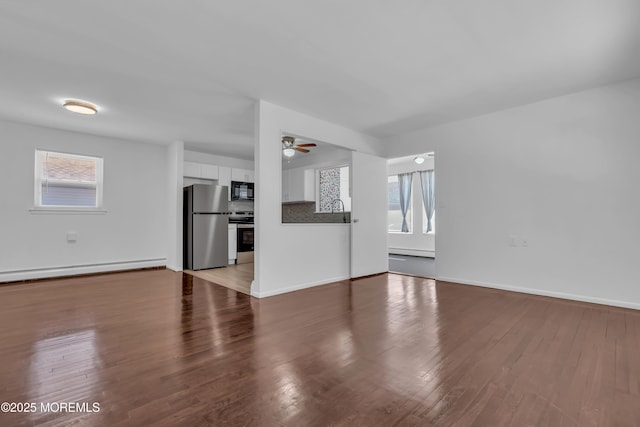 unfurnished living room featuring baseboard heating, plenty of natural light, a ceiling fan, and wood finished floors