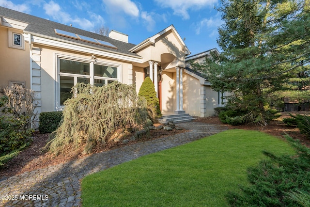 view of front of property with stucco siding, roof with shingles, solar panels, and a front yard