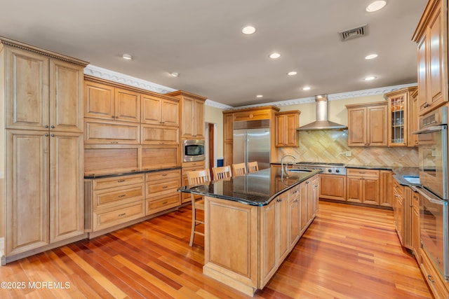 kitchen featuring visible vents, wall chimney range hood, built in appliances, an island with sink, and a kitchen bar