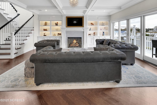 living room featuring stairway, built in shelves, coffered ceiling, and a warm lit fireplace