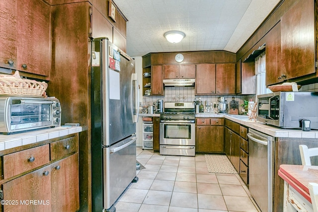 kitchen featuring tile countertops, light tile patterned floors, under cabinet range hood, appliances with stainless steel finishes, and tasteful backsplash