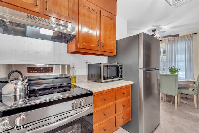 kitchen with backsplash, under cabinet range hood, light countertops, brown cabinets, and appliances with stainless steel finishes