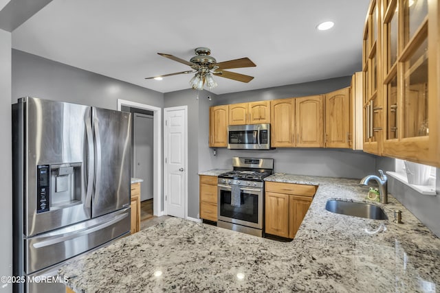 kitchen with a sink, light stone counters, stainless steel appliances, glass insert cabinets, and ceiling fan