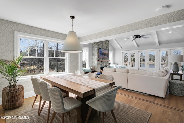 dining area with a healthy amount of sunlight, wood finished floors, wallpapered walls, vaulted ceiling with beams, and a stone fireplace