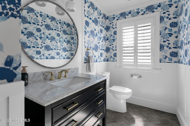 bathroom featuring a wainscoted wall, toilet, crown molding, wallpapered walls, and vanity