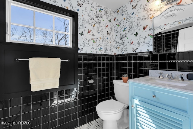 half bathroom featuring a wainscoted wall, toilet, tile walls, wallpapered walls, and vanity