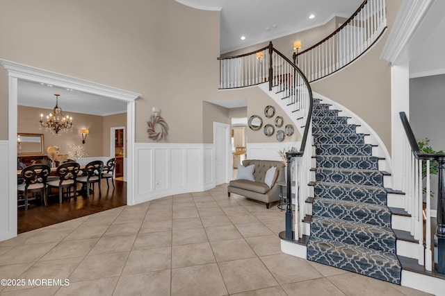 stairs with tile patterned flooring, crown molding, wainscoting, a towering ceiling, and a notable chandelier