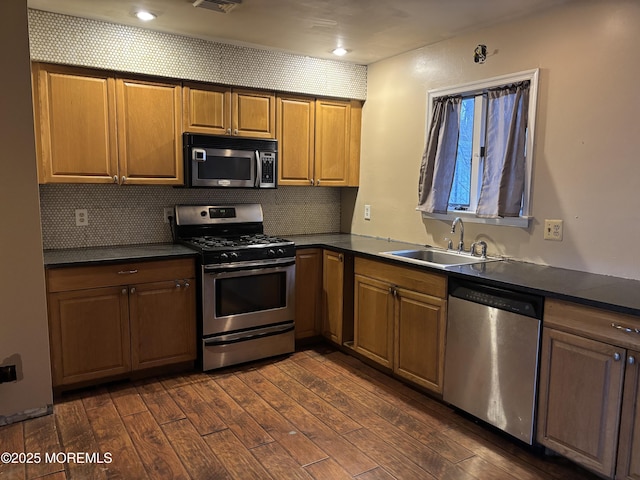 kitchen with dark wood finished floors, a sink, appliances with stainless steel finishes, dark countertops, and backsplash