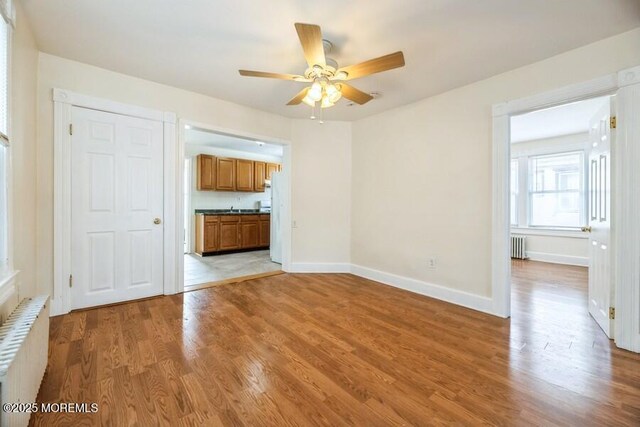 unfurnished living room featuring radiator heating unit, radiator, baseboards, light wood-type flooring, and a ceiling fan