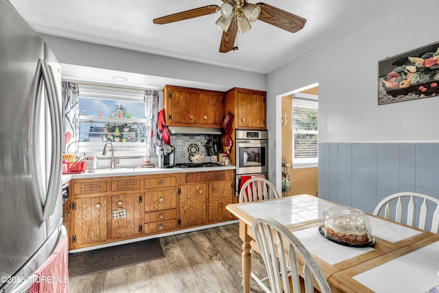 kitchen featuring wood finished floors, brown cabinetry, a wainscoted wall, a sink, and stainless steel appliances