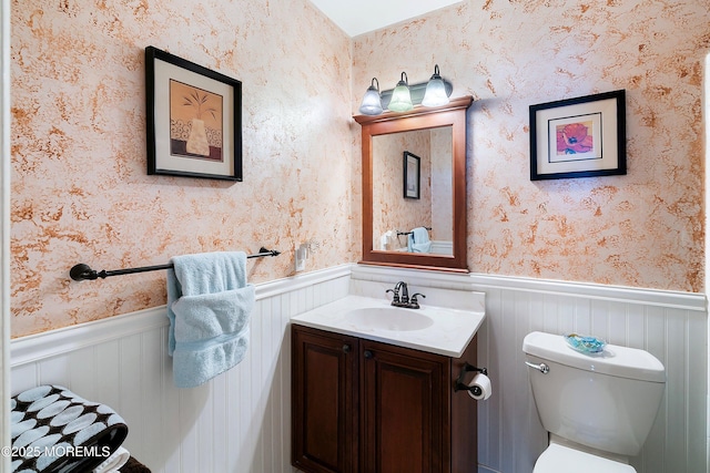 half bath with wainscoting, wallpapered walls, vanity, and toilet