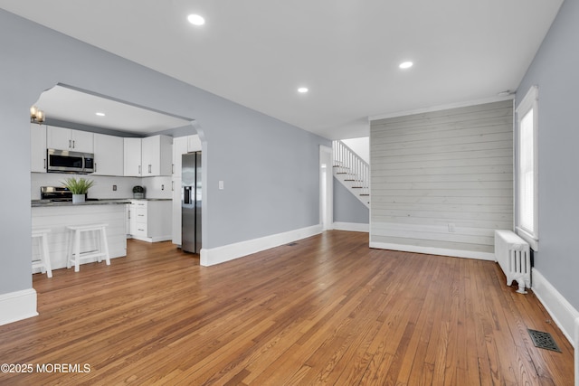 unfurnished living room with stairway, radiator, visible vents, arched walkways, and light wood-type flooring