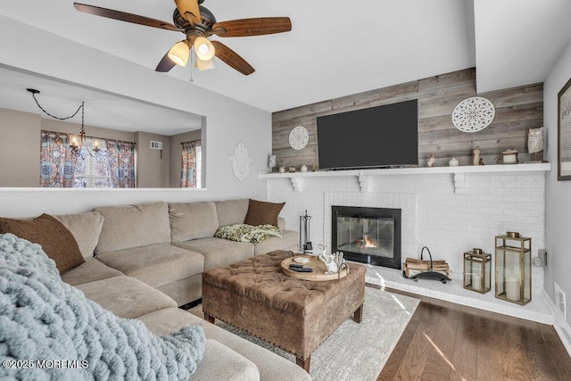 living area with visible vents, ceiling fan with notable chandelier, wood finished floors, wooden walls, and a fireplace