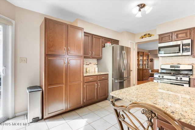 kitchen with light stone counters, brown cabinetry, light tile patterned flooring, appliances with stainless steel finishes, and tasteful backsplash