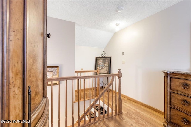 hall with an upstairs landing, lofted ceiling, light wood-style flooring, a textured ceiling, and baseboards