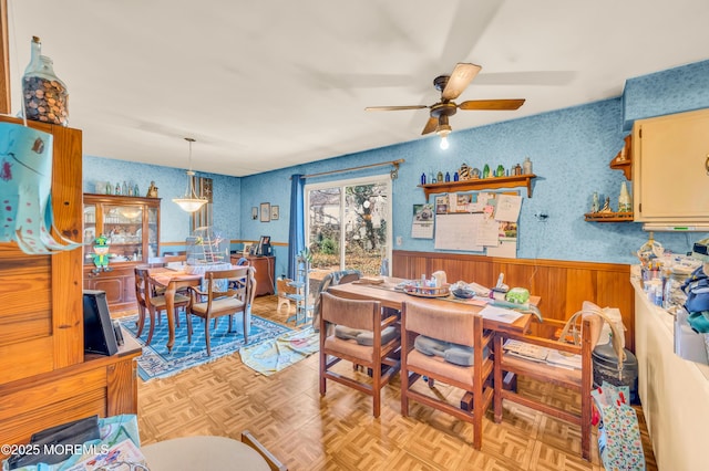 dining space with wallpapered walls, a ceiling fan, and a wainscoted wall