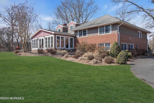 rear view of property with driveway, brick siding, a chimney, and a lawn