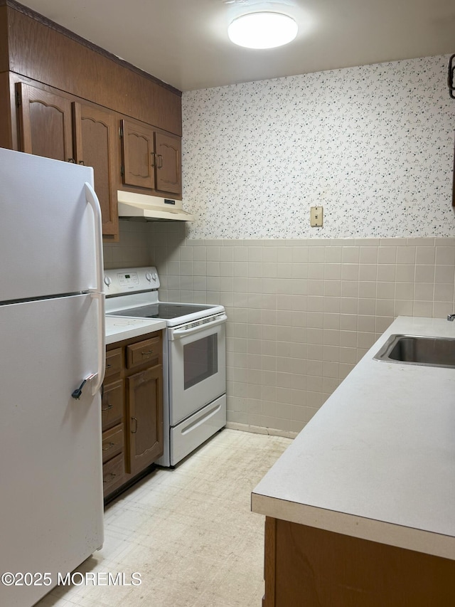 kitchen with a wainscoted wall, under cabinet range hood, white appliances, tile walls, and wallpapered walls