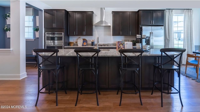 kitchen featuring light stone counters, wood finished floors, a kitchen island with sink, appliances with stainless steel finishes, and wall chimney exhaust hood