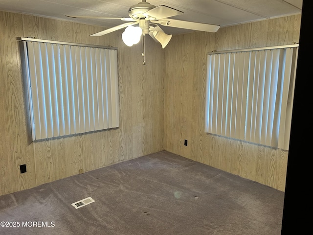 empty room with a ceiling fan, visible vents, carpet floors, and wood walls