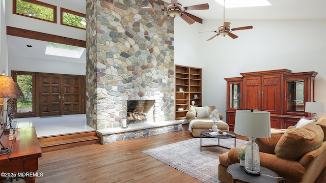 living area with a skylight, high vaulted ceiling, ceiling fan, a stone fireplace, and wood finished floors