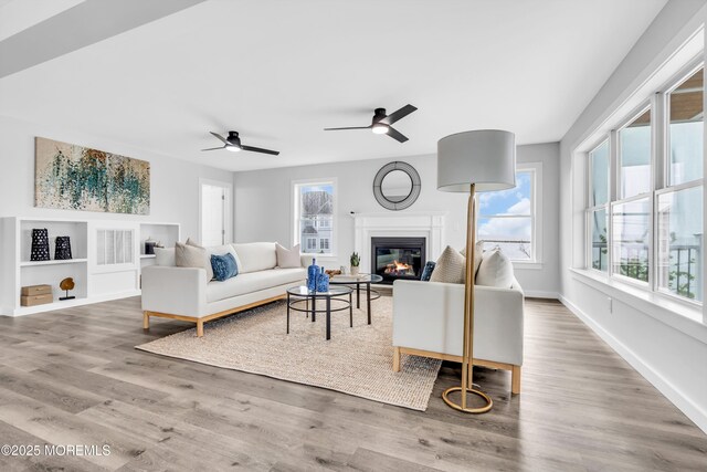 living area featuring ceiling fan, a glass covered fireplace, wood finished floors, built in shelves, and baseboards