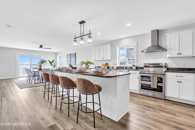 kitchen with range with two ovens, wall chimney exhaust hood, a kitchen island, a kitchen bar, and light wood finished floors