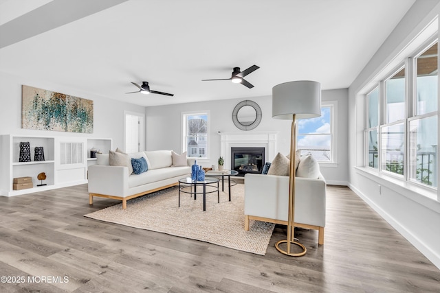 living area featuring ceiling fan, wood finished floors, a glass covered fireplace, baseboards, and built in shelves
