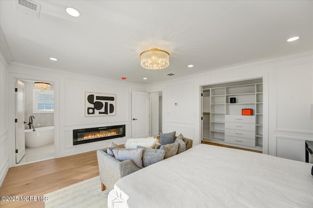bedroom with a decorative wall, crown molding, light wood-style floors, a glass covered fireplace, and a notable chandelier