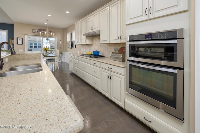 kitchen featuring a notable chandelier, a sink, appliances with stainless steel finishes, under cabinet range hood, and dark wood-style flooring