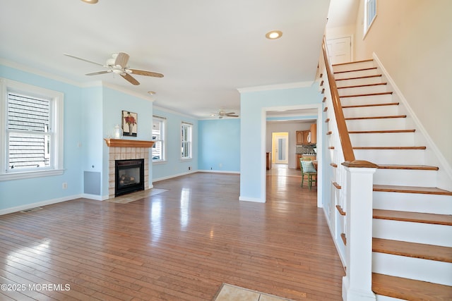 unfurnished living room featuring ceiling fan, crown molding, stairs, a tiled fireplace, and hardwood / wood-style flooring