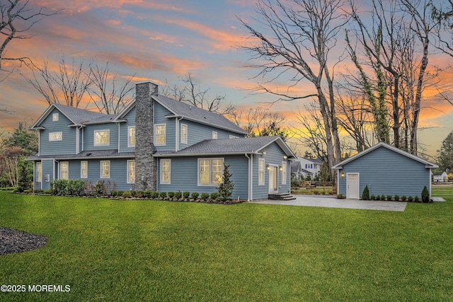 back of house at dusk featuring an outdoor structure, a yard, a shingled roof, and a chimney
