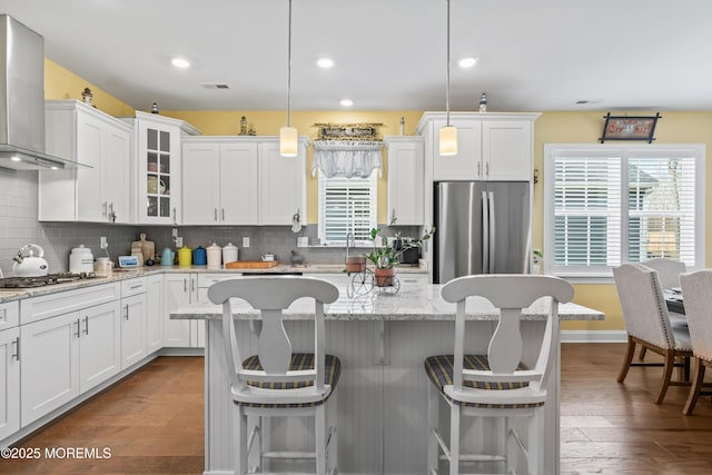 kitchen with glass insert cabinets, wall chimney range hood, white cabinetry, a breakfast bar, and appliances with stainless steel finishes