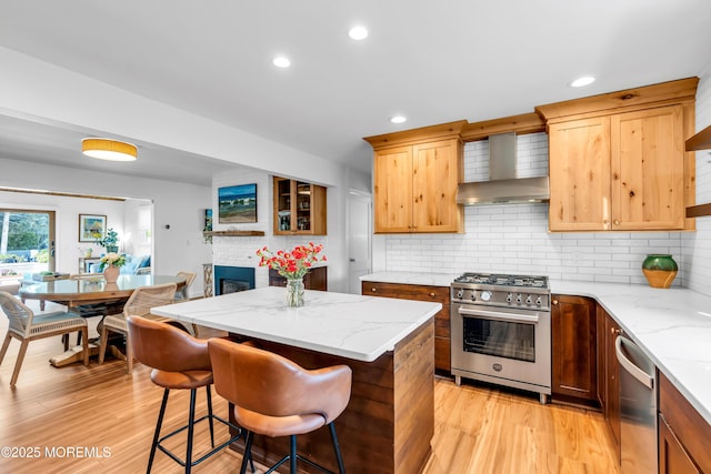 kitchen featuring wall chimney range hood, appliances with stainless steel finishes, a kitchen island, light wood finished floors, and recessed lighting