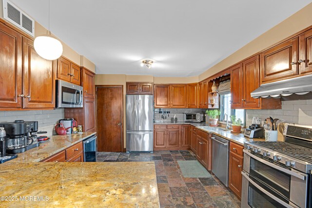 kitchen with stone finish flooring, a sink, appliances with stainless steel finishes, under cabinet range hood, and visible vents