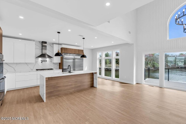 kitchen with hanging light fixtures, wall chimney exhaust hood, modern cabinets, light wood-style floors, and stainless steel appliances