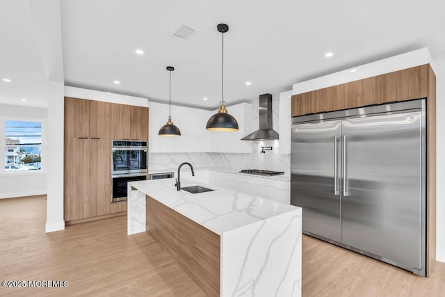 kitchen featuring wall chimney range hood, modern cabinets, brown cabinetry, stainless steel appliances, and a sink