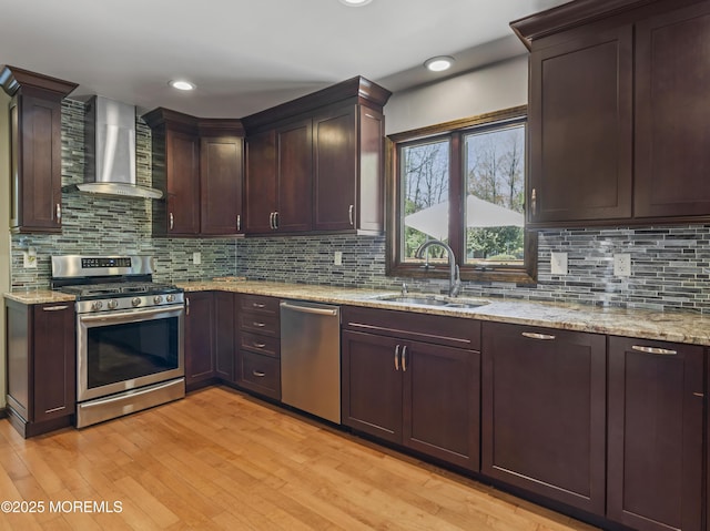 kitchen featuring wall chimney range hood, light stone counters, a sink, stainless steel appliances, and light wood-type flooring