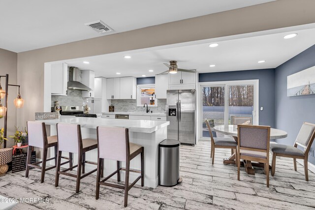 kitchen featuring wall chimney exhaust hood, a peninsula, appliances with stainless steel finishes, light countertops, and a sink