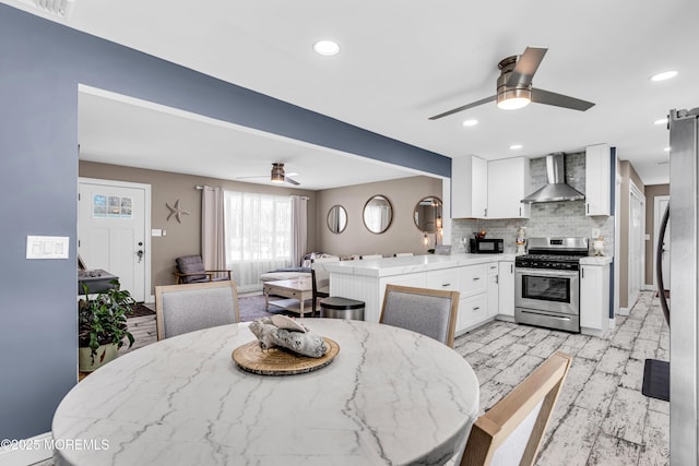 kitchen with wall chimney exhaust hood, appliances with stainless steel finishes, a ceiling fan, white cabinetry, and backsplash