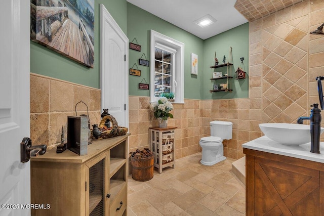 bathroom with tile walls, toilet, vanity, wainscoting, and stone finish flooring